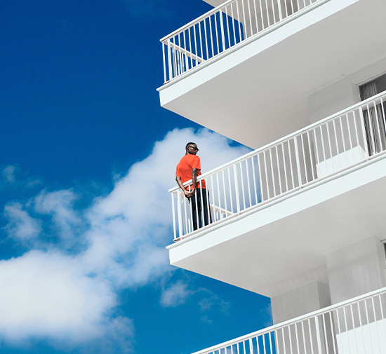 Hombre de pie en el balcón de un hotel con cielo azul y nubes blancas de fondo