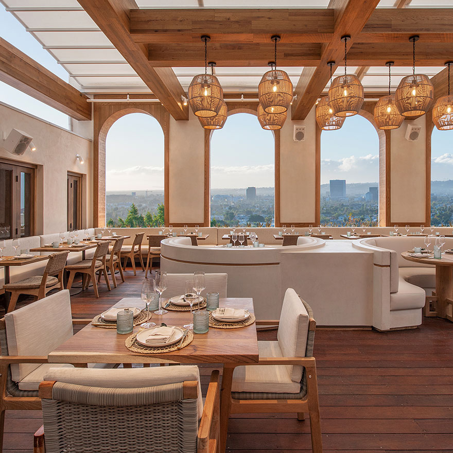 Comedor de madera clara y color crema con lámparas colgantes de ratán, pequeña mesa cuadrada de madera con sillas de comedor color crema en primer plano, y grandes cabinas color crema al fondo con ventanas curvas con vistas al paisaje urbano de Los Ángeles.