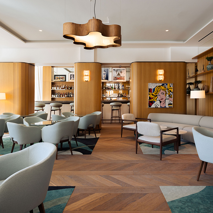 A lounge with modern gray-upholstered furnishings, wooden floors and lighter, wooden-panel walls, with sconces and a pop-art painting of a woman, and a bar in the background.