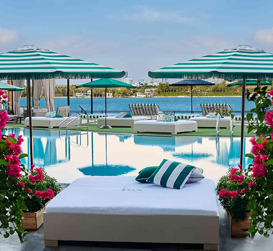 Cama junto a la piscina con almohadas a rayas verdes y blancas, rodeada de flores rosas, con la bahía de Biscayne al fondo.