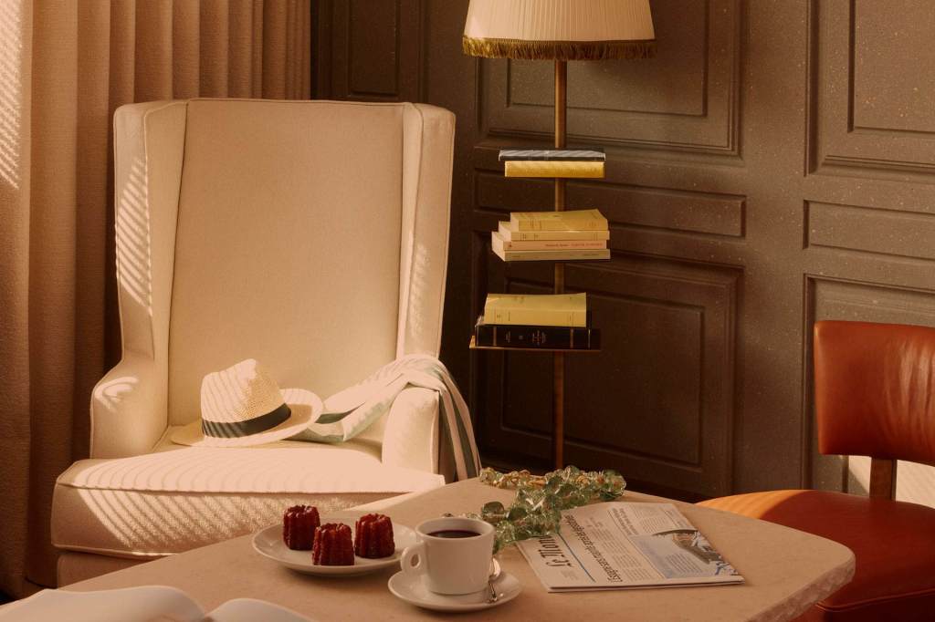 Hotel guestroom with a white chair with a hat, and lamp with bookcase