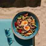 Salad with hardboiled eggs in a blue bowl sitting on a blue lounge chair with a pair of sunglasses