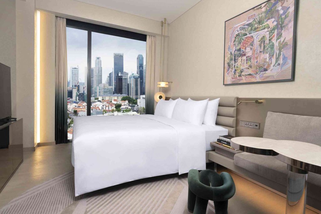 A spacious hotel room with a bed, a TV, a painting and a stunning city view from the window.