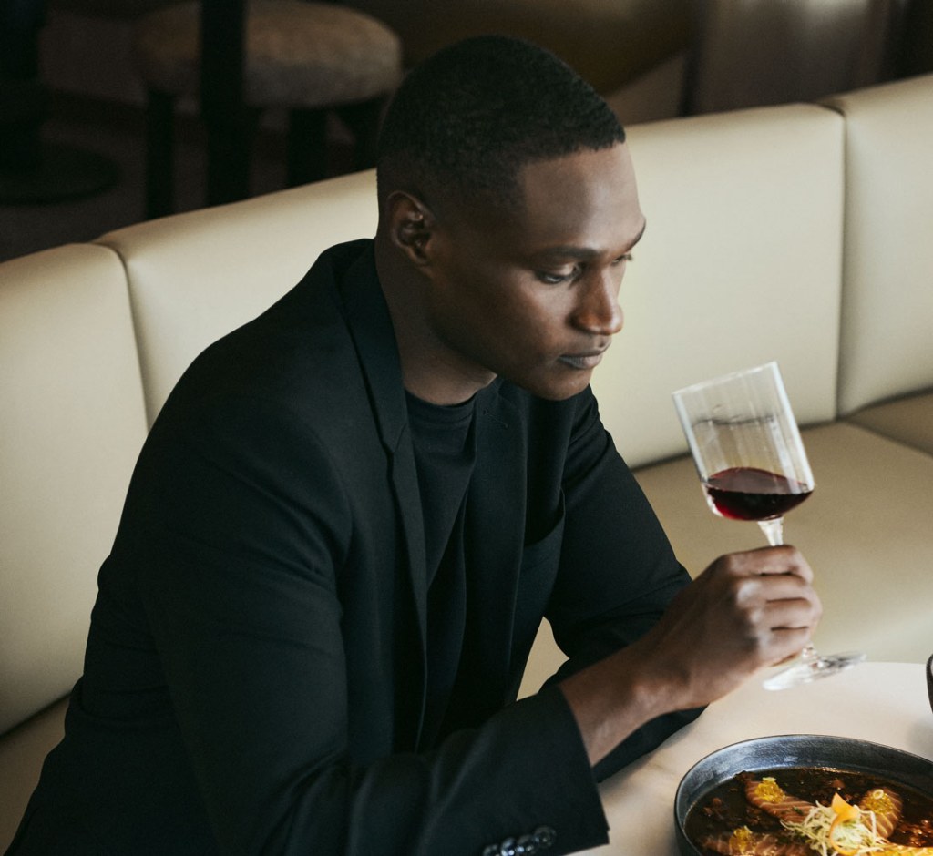 Hombre vestido elegantemente con un traje negro sentado en una mesa de comedor, con una copa de vino tinto en la mano