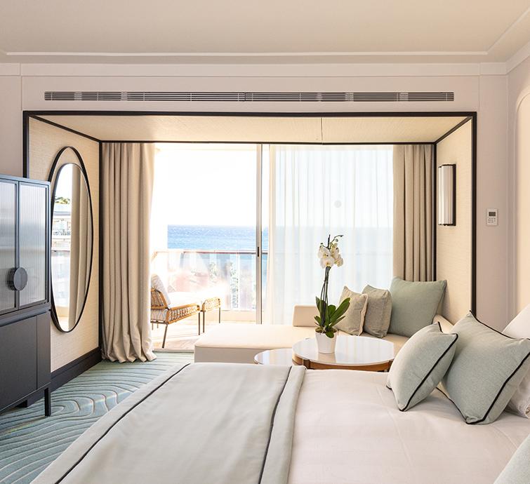 Cama en habitación de hotel con ropa de cama verde claro, un gran espejo ovalado en la pared, junto a una zona de estar y un balcón con vistas al mar 