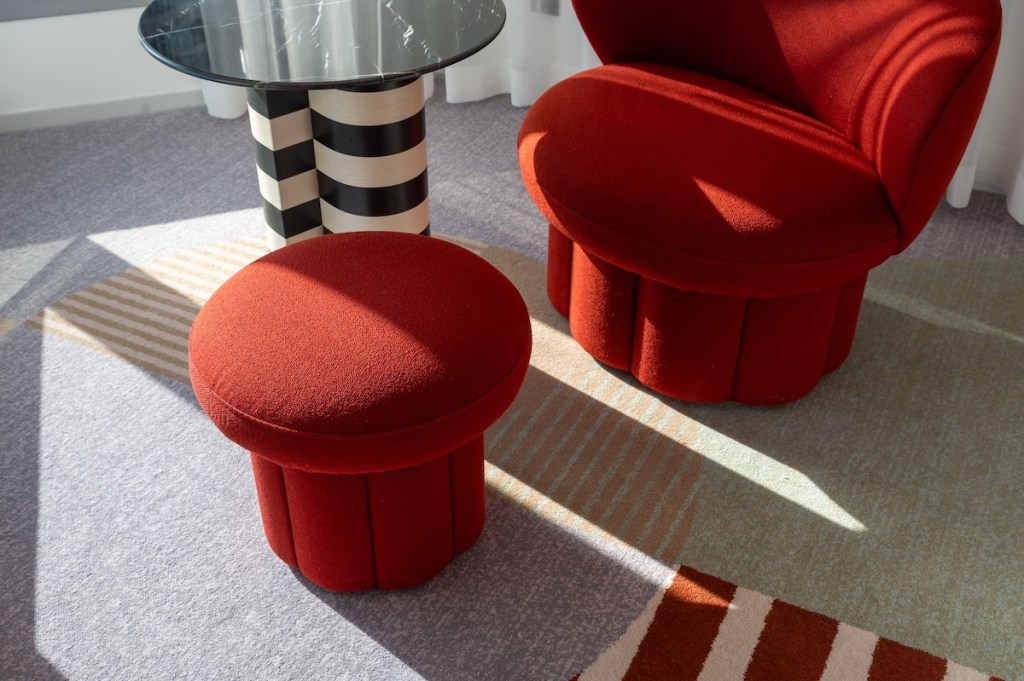 Red and white striped rug in a room with a red chair, red stool, small black and white table, and white curtains covering a window