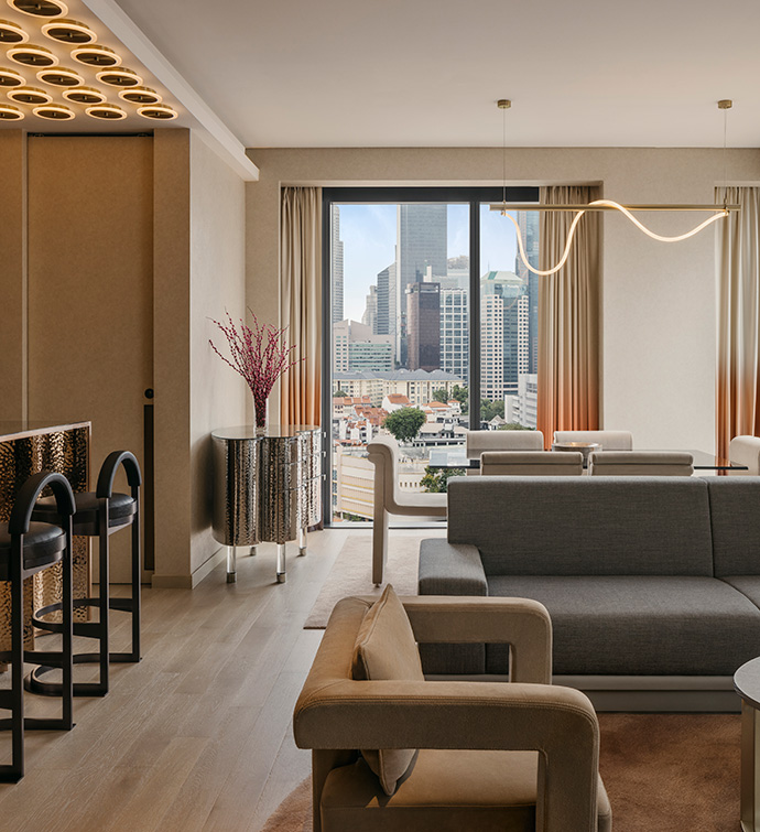 Living room of suite at Mondrian Singapore Duxton with modern seating in neutral tones  to the right, black barstools under golden lighting, and a tube of neon lighting hanging in the background in front of a view of the city. 