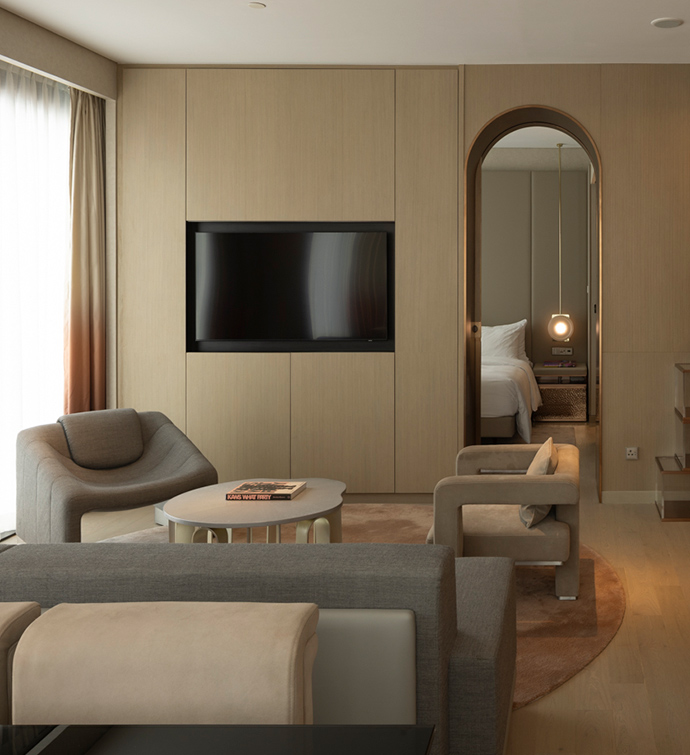 Living room of suite at Mondrian Singapore Duxton with modern seating upholstered in gray hues facing a television and an arched doorway through which a bed is visible