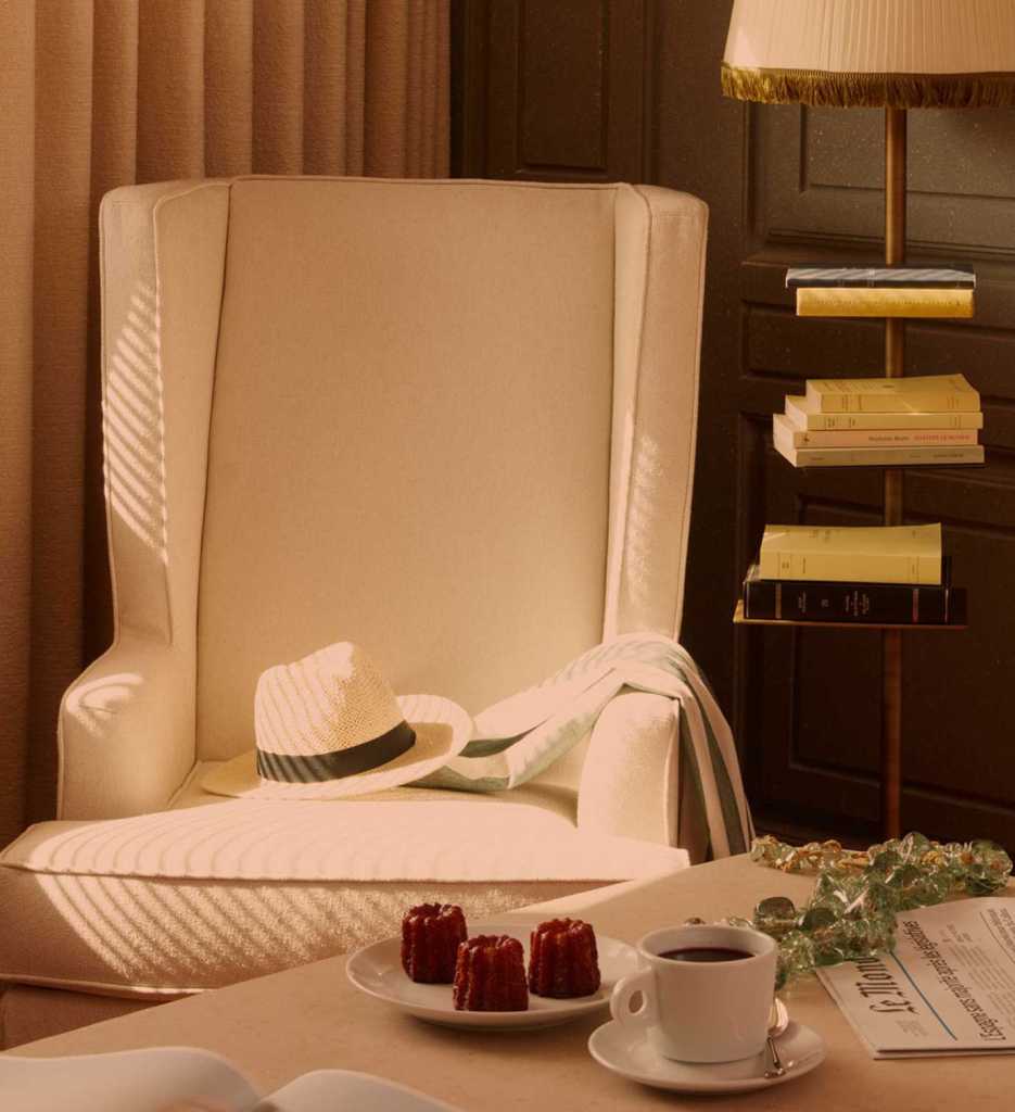 High back arm chair upholstered in cream, where someone has placed a white straw hat, in front of a coffee table with coffee and pastries