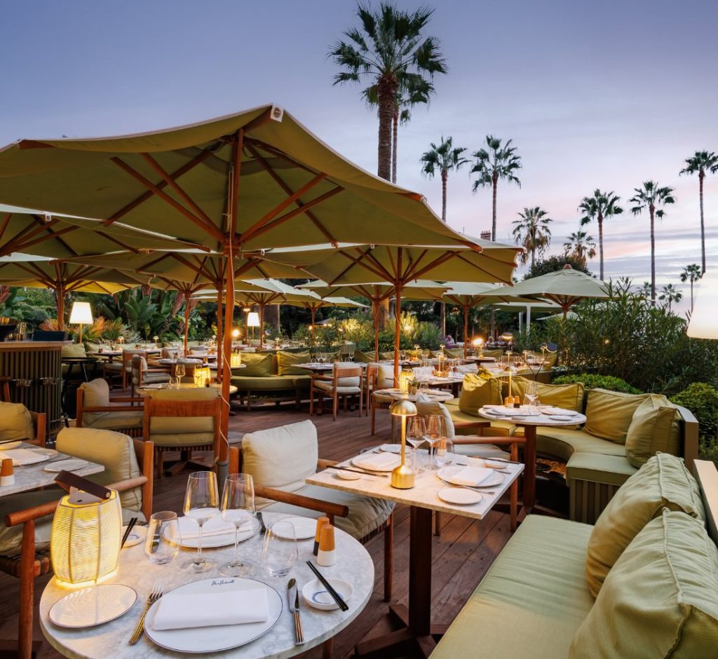 An outdoor dining patio in Cannes at dusk, with yellow umbrellas and palm trees in the distance