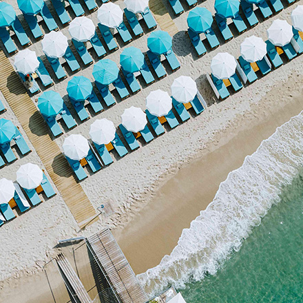 Aerial image of a sandy beach and turquoise water with rows of white and turquoise umbrellas and chaise lounge chairs.