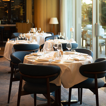 Elegant table with cream-colored tablecloth, dark blue upholstered and wooden dining chairs, and wine glasses and tableware placed on the table. Similar round tables in the background. 