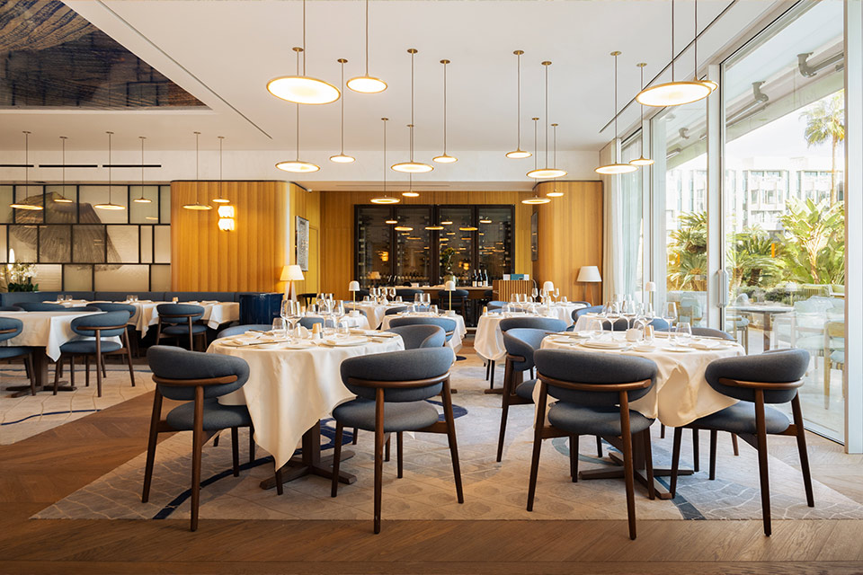 Grande salle à manger de restaurant avec de petites tables aux nappes couleur crème, des chaises en bois bleu foncé, des lumières suspendues et un plancher en bois avec un grand tapis.