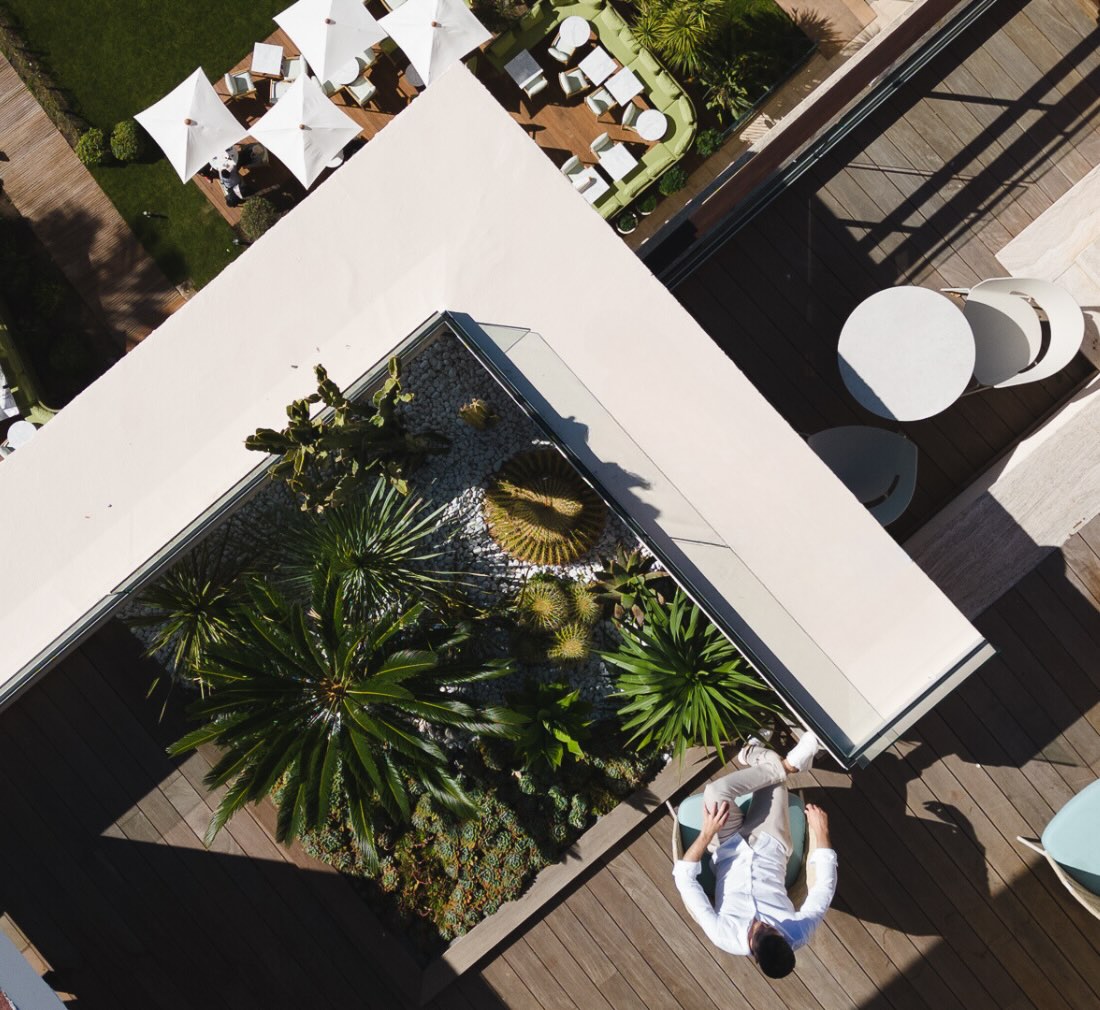 Una terraza elevada captada desde arriba durante el día, que muestra a un hombre sentado en una silla junto a un pequeño jardín de palmeras. En el suelo, a lo lejos, hay un patio con mesas y sombrillas.