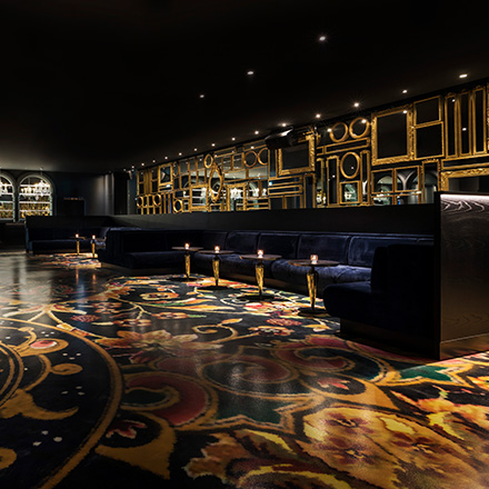 Dark lounge space with velvet blue sofas and small cocktail tables with an intricate gold and colorfully designed floor.