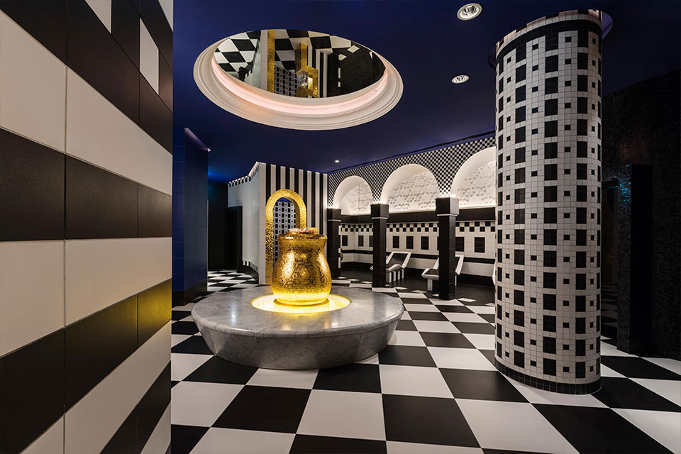 Spa Mondrian Doha avec des sols à damier noir et blanc, un plafond bleu avec un miroir et un grand coin salon en or et en marbre.