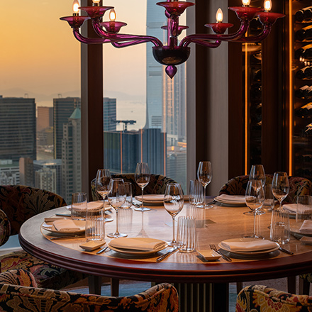 Warmly lit dining room with round wooden dining table with plates, silverware, and wine glasses on top. A traditional chandelier is hanging above the table and large floor-to-ceiling windows show a cityscape in the background.