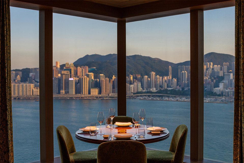 Small round dining table with three cushioned dining chairs in front of large floor-to-ceiling windows looking out to a cityscape with mountains in the background.