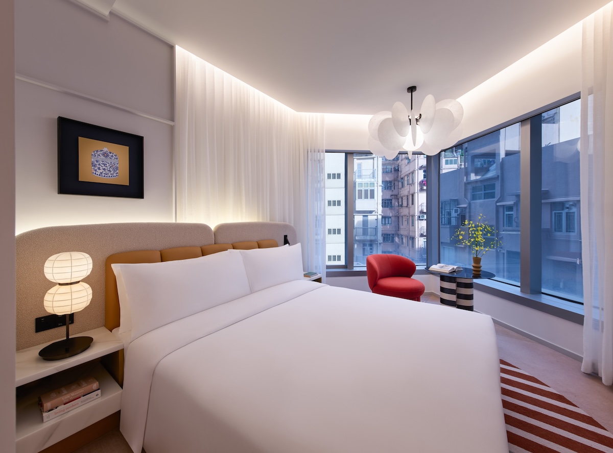 Large white bed with grey and yellow headboard, with white curtains, window looking out to the city, and small black and white table with red lounge chair