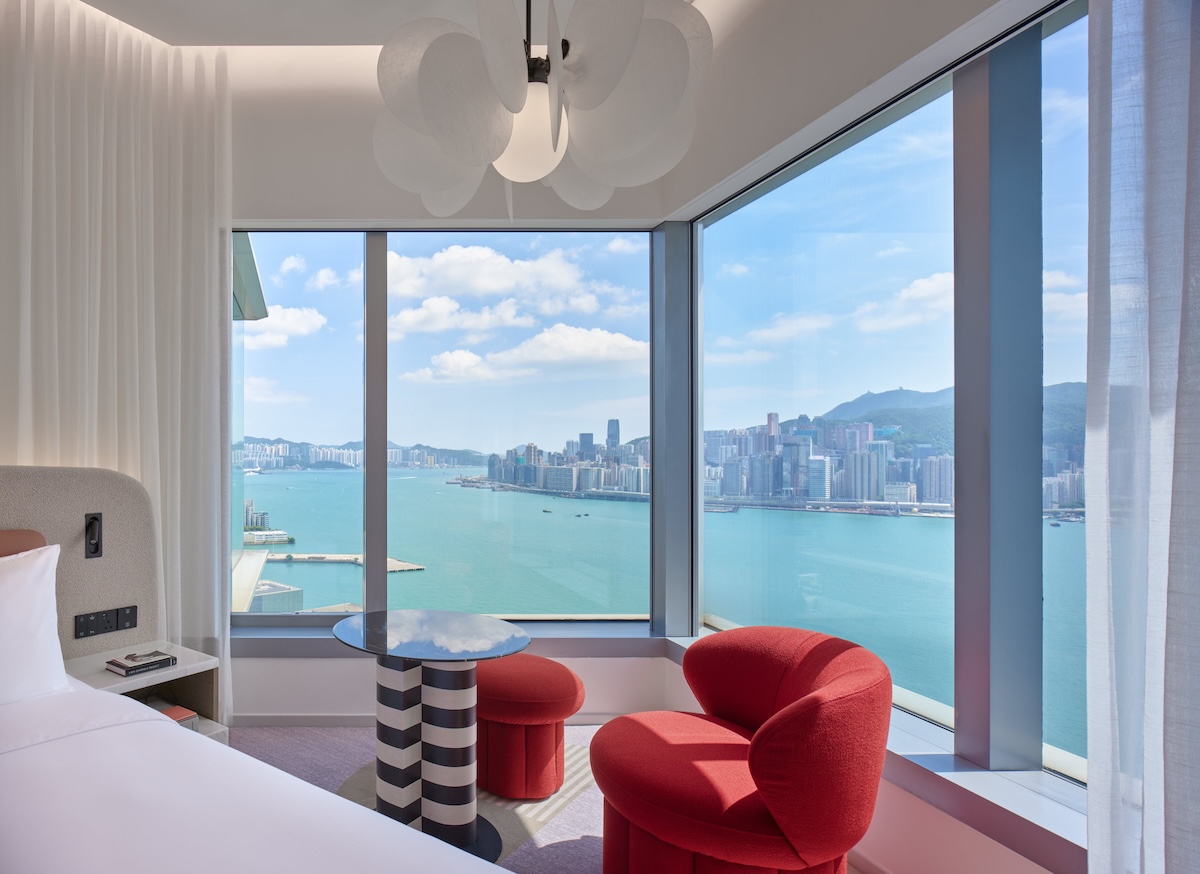 Side of white bed with window view of city and small table and red chair in the corner