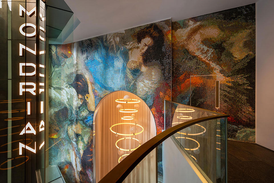 Hotel lobby view from a higher floor, with an illuminated Mondrian vertical logo on the left, a modern lit chandelier hanging from the ceiling, and a colorful mosaic mural on the wall.