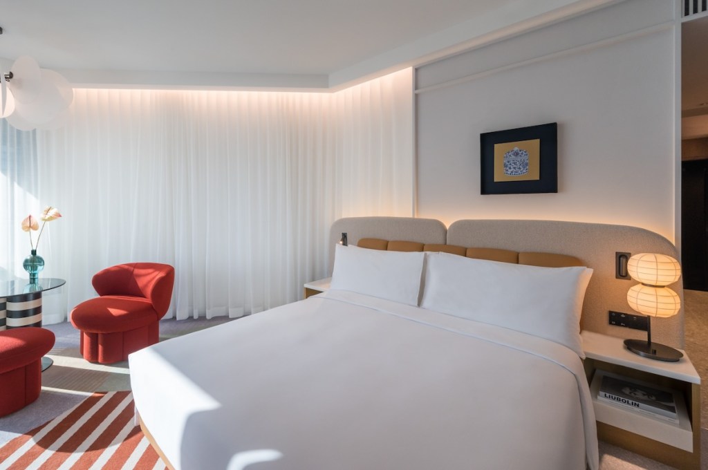 Large white bed with grey and brown headboard at an angle with red striped rug underneath, small table and red chair in the corner