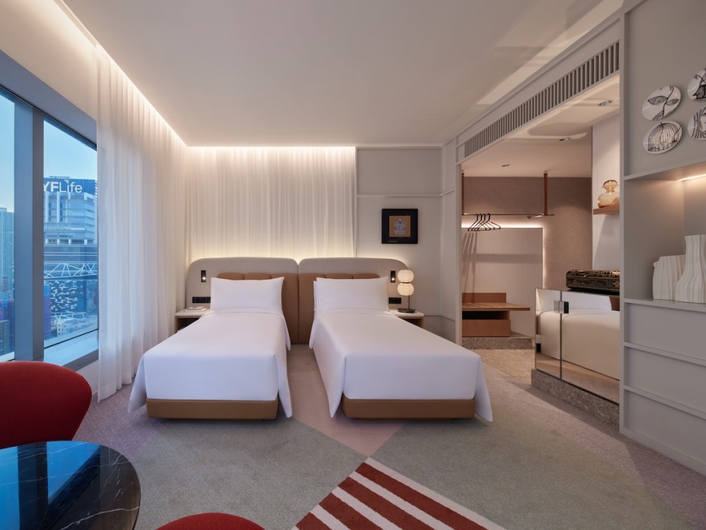 Two white twin beds with a grey and beige shared headboard with white curtains behind it, a geometric patterned carpet, and closet in the corner