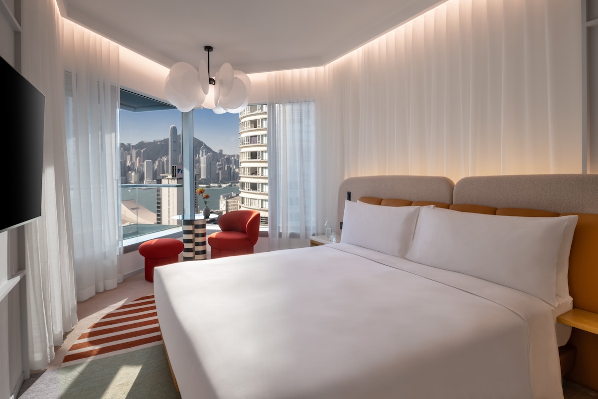 Large white bed with grey headboard, small red chair and black table in the corner, and view of a cityscape out of the large windows