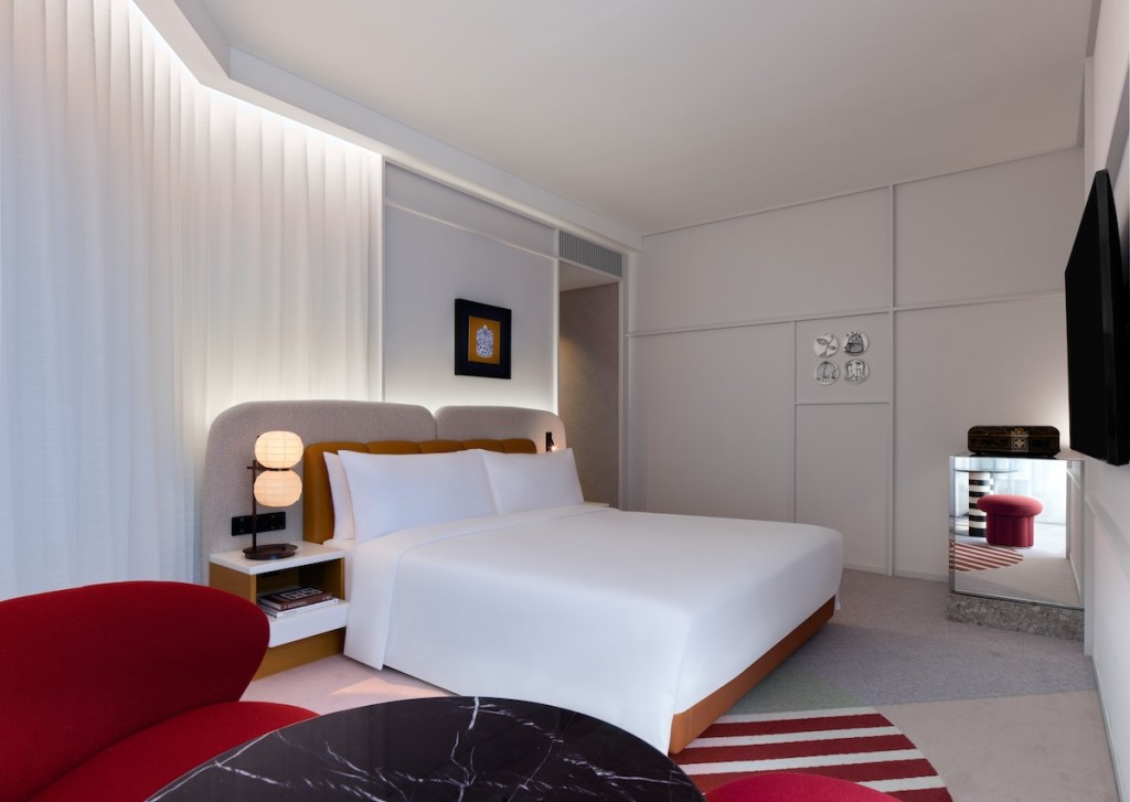 Large white bed with grey and brown headboard at an angle with red striped rug underneath, small table and red chair in the corner