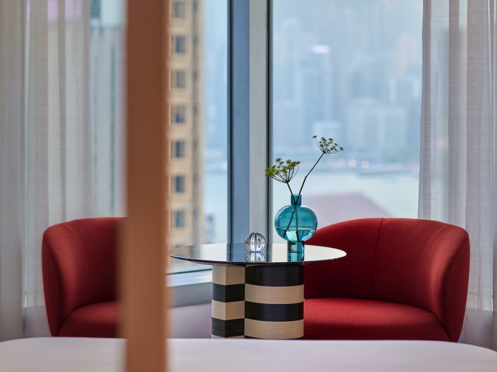 small black and white striped table with glass and book on top and two red chairs nearby and a window looking out to the city