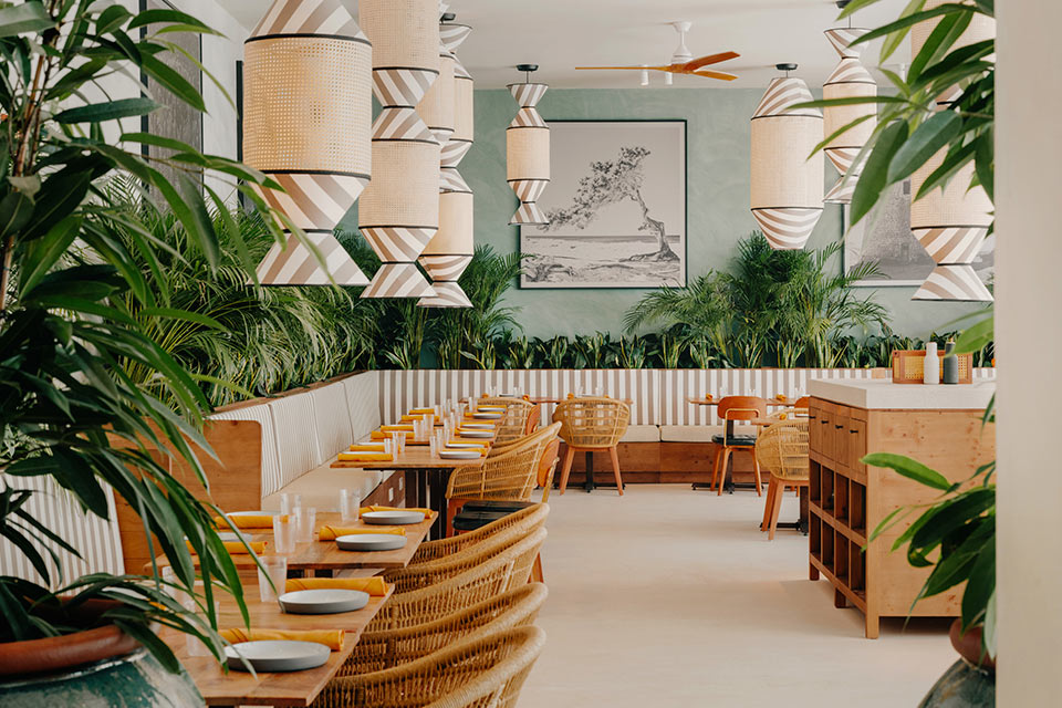 Espacio de comedor con mesas cuadradas de madera, sillas de mimbre, plantas verdes esparcidas por todas partes y lámparas colgantes con diseño de rayas. 