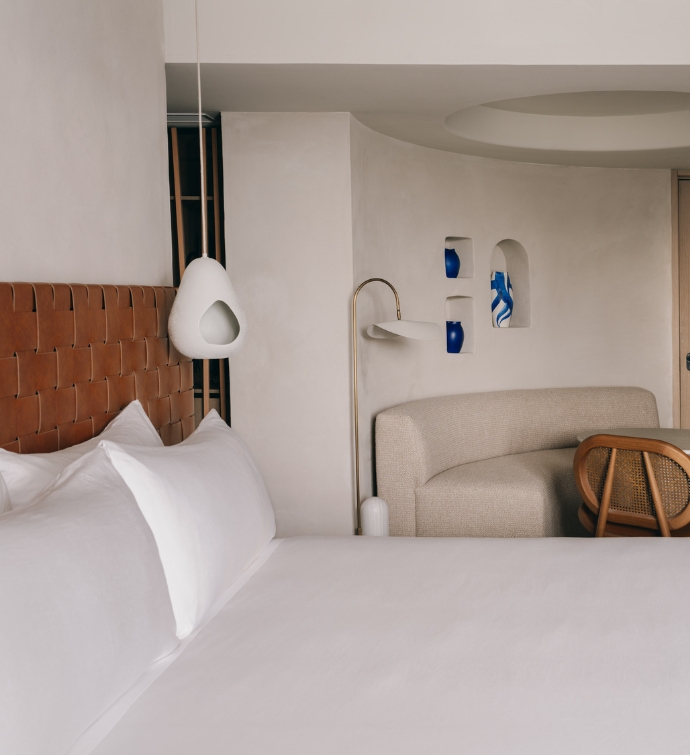 Side of white bed with rust colored headboard and a cream bench and wooden chair in the corner