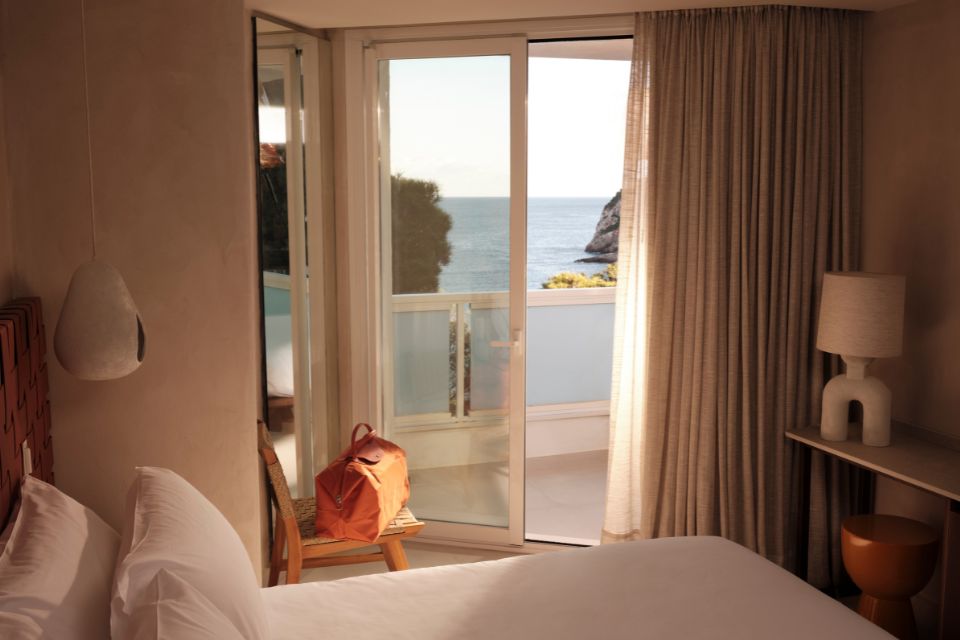 Bedroom with natrual light seeping through the balcony windows, a large white bed in the middle, windows looking out to the sea, and an orange bag sitting on a bench