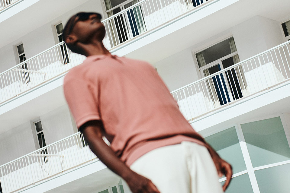 Hombre con gafas de sol, camisa rosa y pantalones blancos mirando hacia el cielo con la fachada blanca de Mondrian Ibiza al fondo.