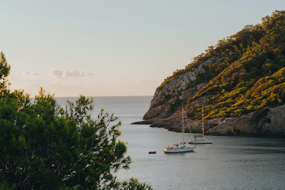 Vista aérea de la bahía de Cala Llonga, con barcos en el agua rodeados de montañas exuberantes.