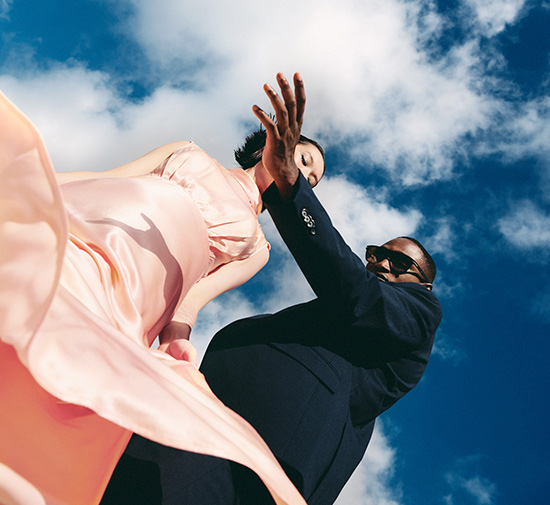 Woman in a pink dress dancing with a man in a suit and sunglasses, with a bright blue sky with clouds in the background.