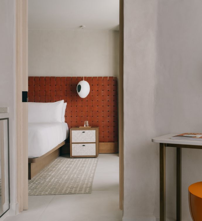 Hotel bedroom view through a door with a white bed, small bedside table, pendant white lamp, and rust colored headboard