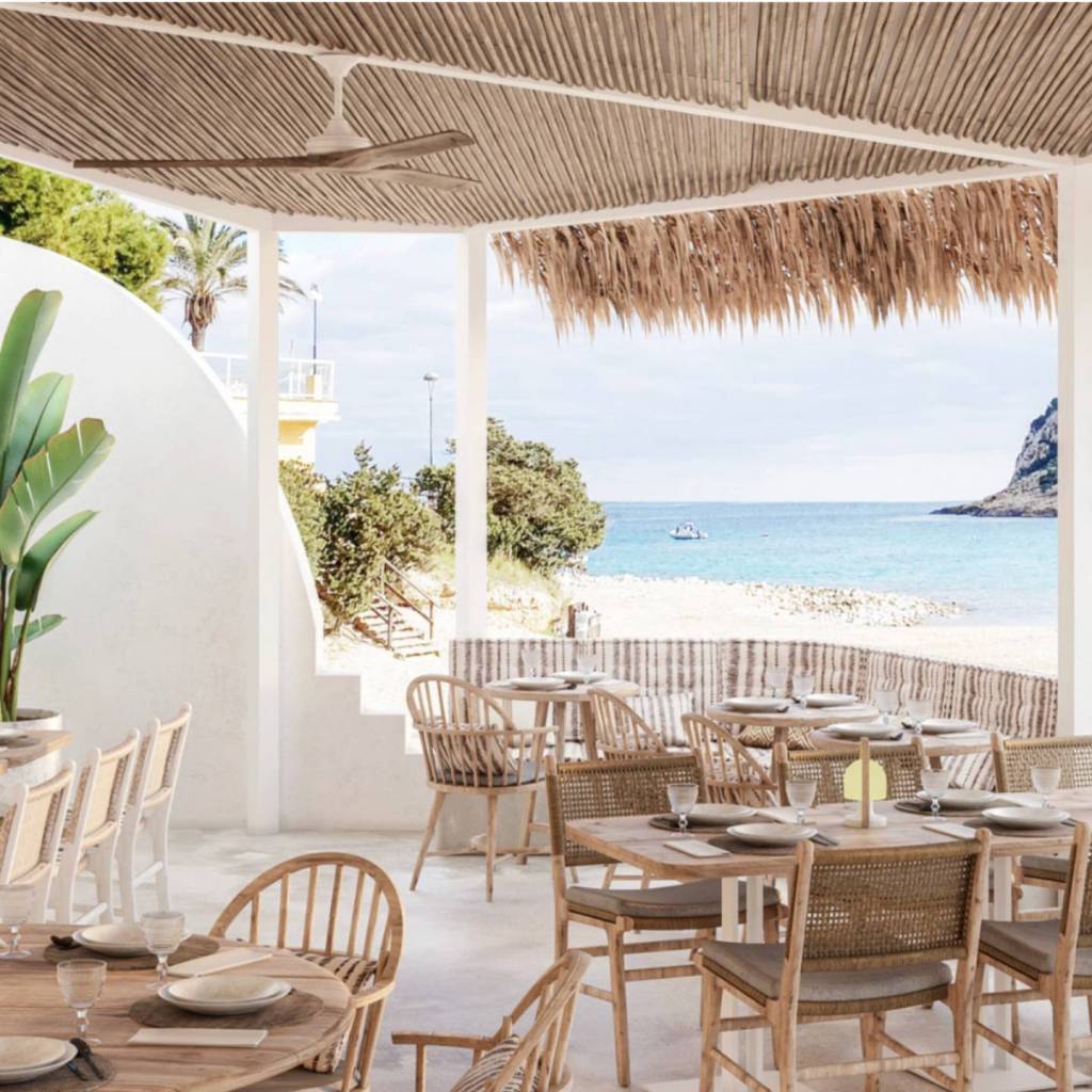 Outdoor patio with light wooden chairs and tables with a beach in the background