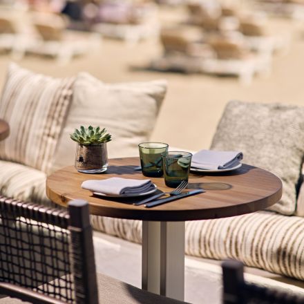 Close up image of wooden round table with small plant, two glasses, and table settings on it with a striped cushion booth and sandy beach in the background