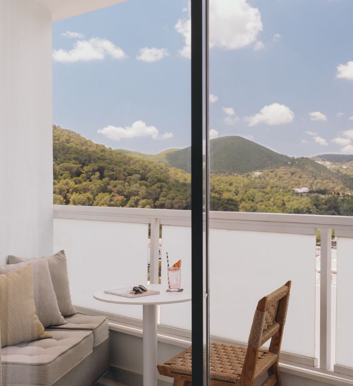 Balcony with cream cushioned bench and pillows, small round white table with drink and sunglasses on top, and a wicker outdoor chair, green mountains and blue sky are in the distance