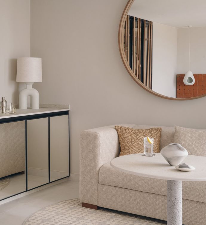 Corner of a cream sofa with small pillow and white round coffee table with a round framed mirror on wall above and a glass hutch with lamp in the corner