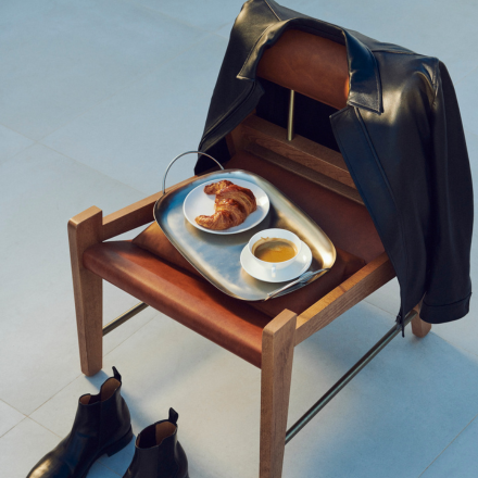 Croissant and coffee on a tray on a wooden chair with leather jacket slung over it and boots on the floor