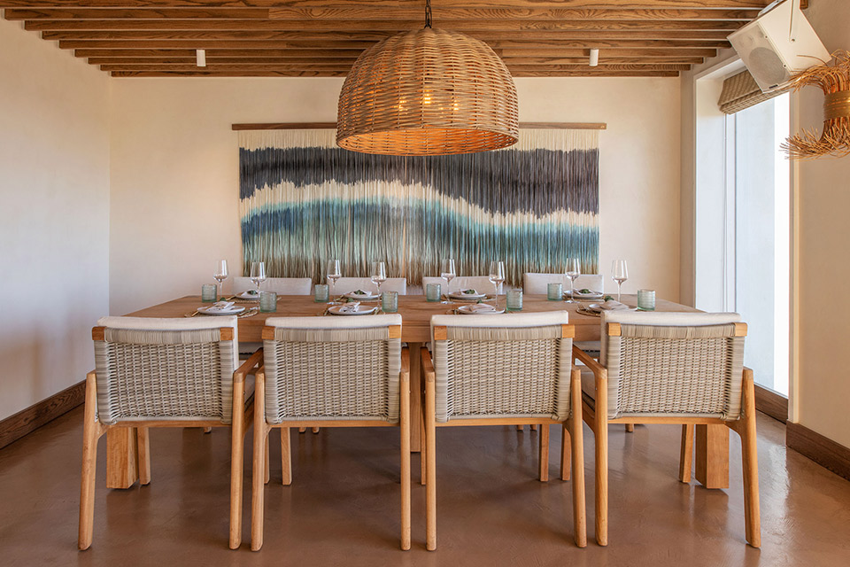 Modern bohemian wooden rectangular dining table with wicker chairs, wicker chandelier, and colorful wall art in the background.