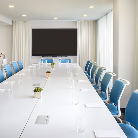 Gran sala de juntas blanca con una larga mesa, sillas azules y televisor.