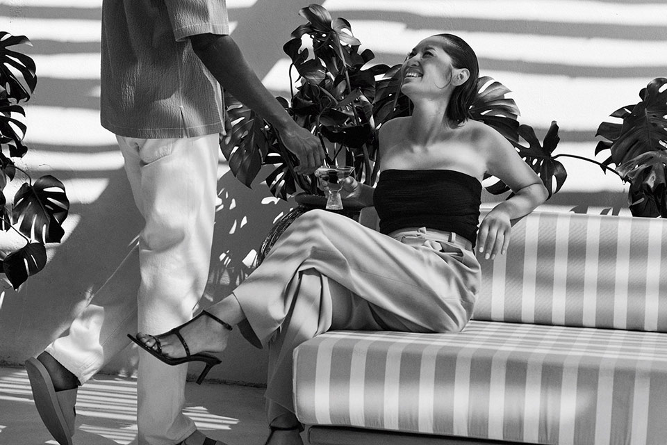 Black and white photo of a woman in a black tube top looking up and smiling at a man, while sitting on a striped couch.