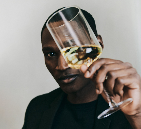 Homme à l'aspect élégant vêtu d'un haut noir, se tenant devant un mur gris, un verre de vin blanc à la main qu'il lève devant son visage en regardant directement la caméra.