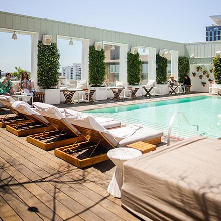 White chaise lounges surround a turquoise pool and pool deck with large windows in the background and greenery throughout.