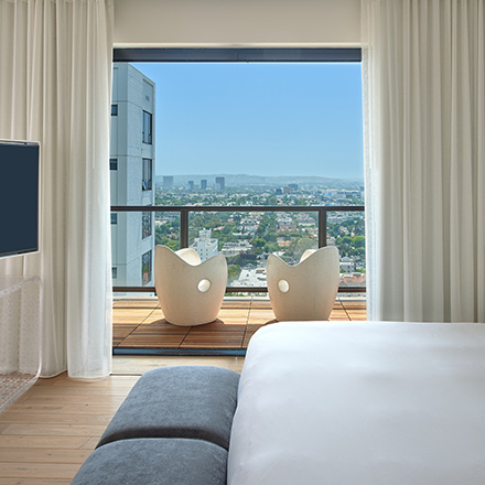 Mondrian Los Angeles guest room balcony with a bed, TV and views of the city.