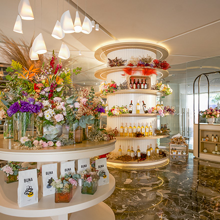 Luminosa tienda de regalos con estanterías curvadas llenas de flores de diferentes colores en jarrones y botellas de vino en estantes al fondo.