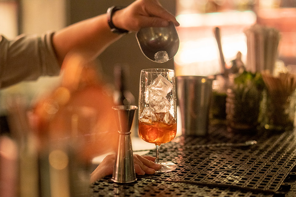 Close-up image of a bartender's arm pouring ice into an orange cocktail in a large clear glass with rocks.